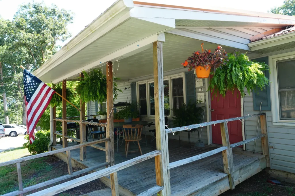 The covered front porch of one of the homes, with hanging ferns, an American flag and a red front door.